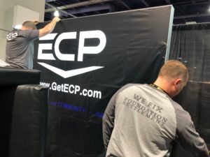 Photo of two men setting up an ECP backdrop for an exhibit space at World of Concrete. The backdrop is all black and says ECP in white letters in the center.